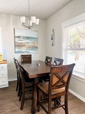 Une salle à manger avec une table et des chaises en bois, un lustre, un tableau et une fenêtre avec des stores.