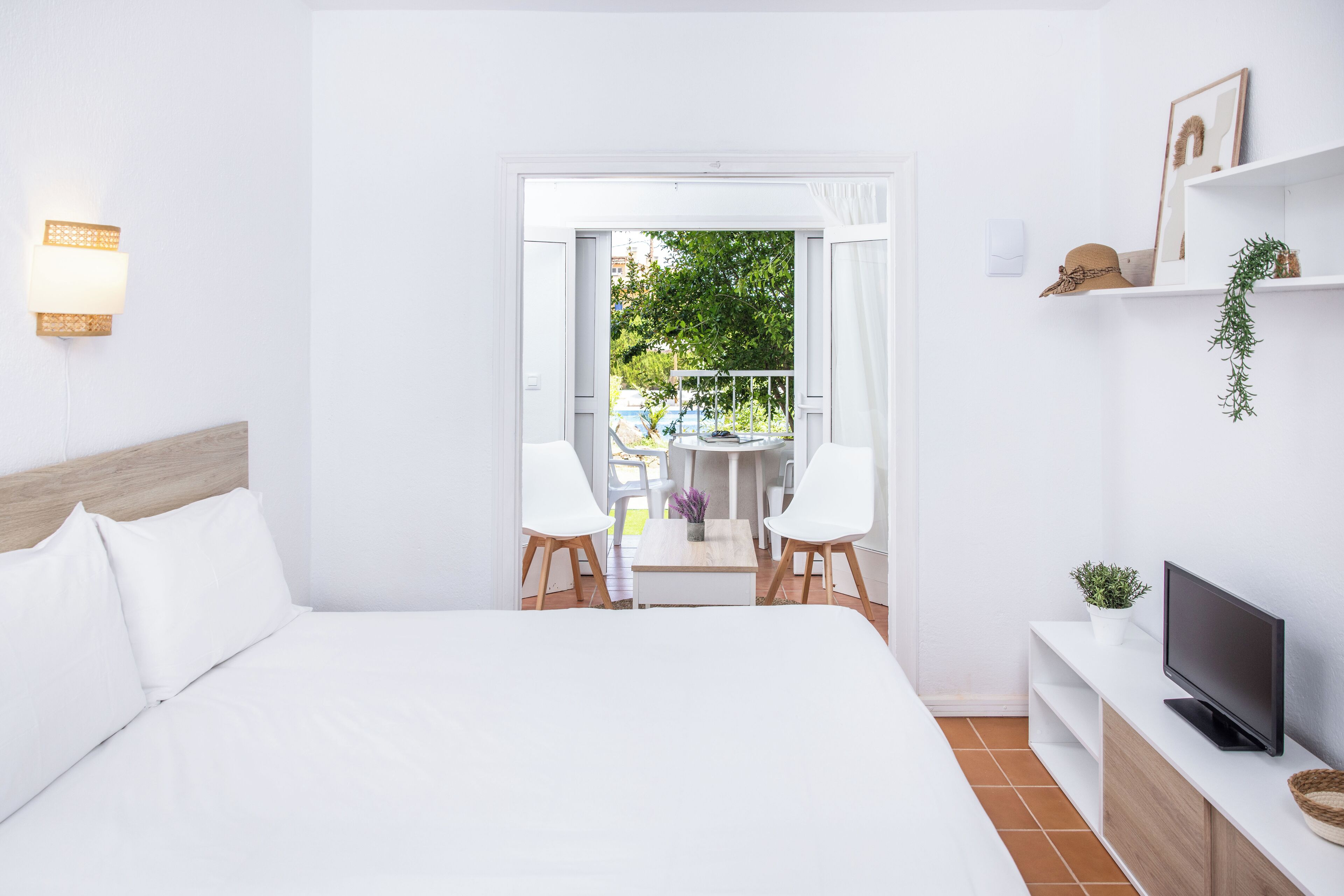 A modern hotel room with a large bed, a TV on a wooden console, a balcony with a table and chairs, and a view of greenery outside.