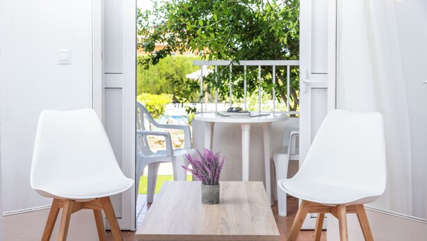 Une pièce moderne avec une table blanche, deux chaises blanches, une petite plante en pot et une vue sur un jardin par une porte ouverte.