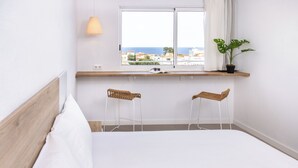 Une chambre avec un lit, deux chaises, une fenêtre donnant sur la mer, une plante et une étagère avec une lampe.