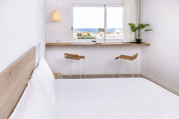 A bedroom with a bed, two chairs, a window with a view of the sea, a plant, and a shelf with a lamp. - RVApartaments Escala Brava (L'Escala)