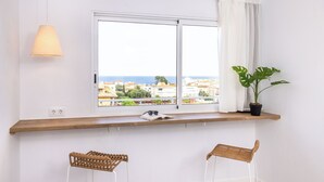 Une pièce avec une grande fenêtre, une étagère en bois sur laquelle se trouvent un livre et une paire de lunettes de soleil, une plante en pot et deux chaises en osier.
