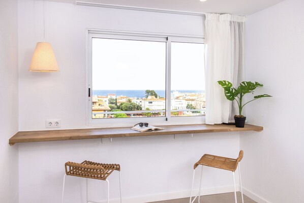 A room with a large window, a wooden shelf with a book and sunglasses, a potted plant, and two wicker chairs. - RVApartaments Escala Brava (L'Escala)