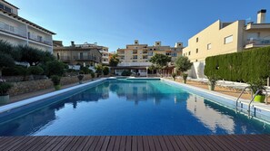 A swimming pool with a wooden deck in the foreground, surrounded by multi-story buildings with balconies and a stone wall. - RVApartaments Escala Brava (L'Escala)