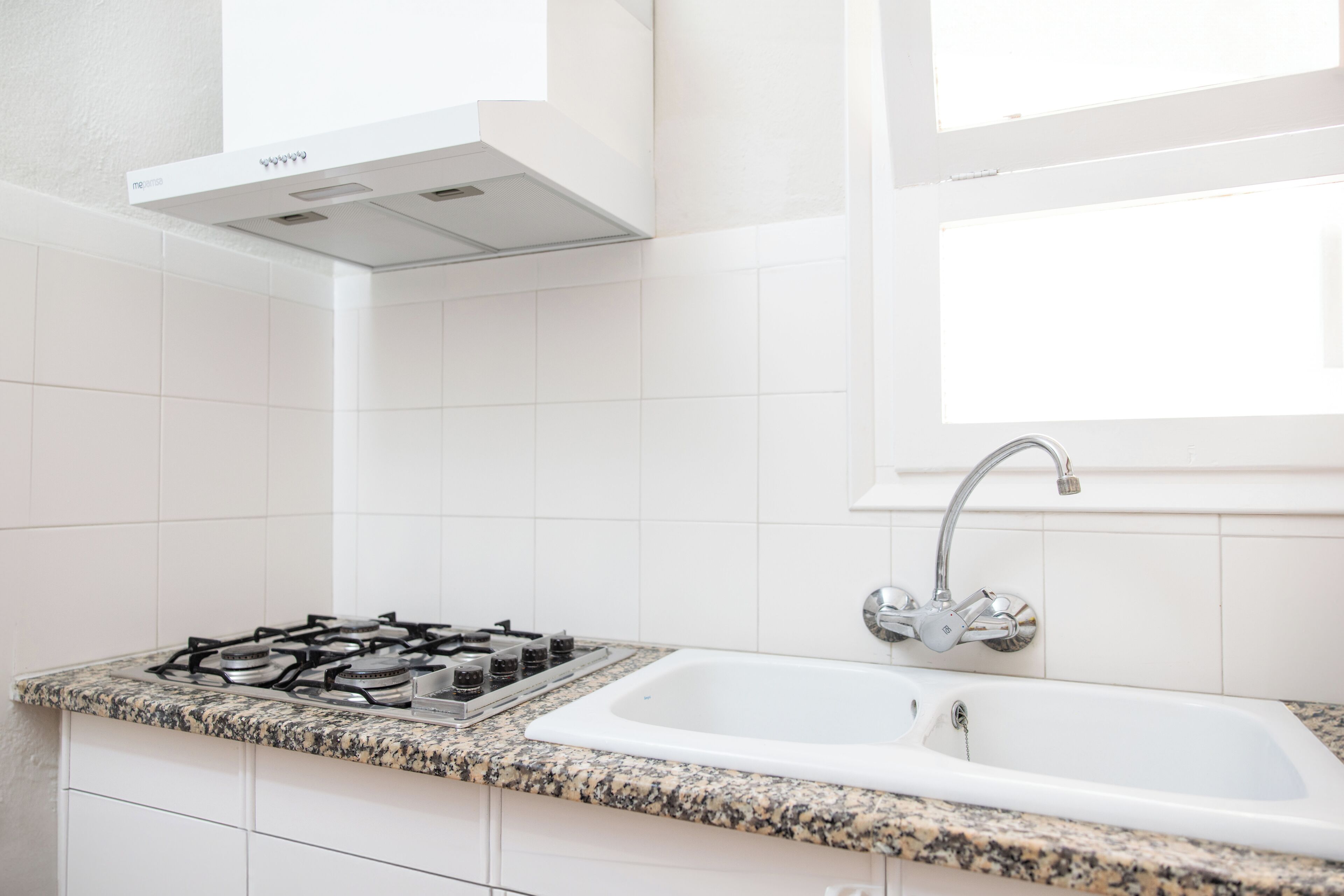 A kitchen with white tiles, a gas stove, a sink, and a window above the sink.