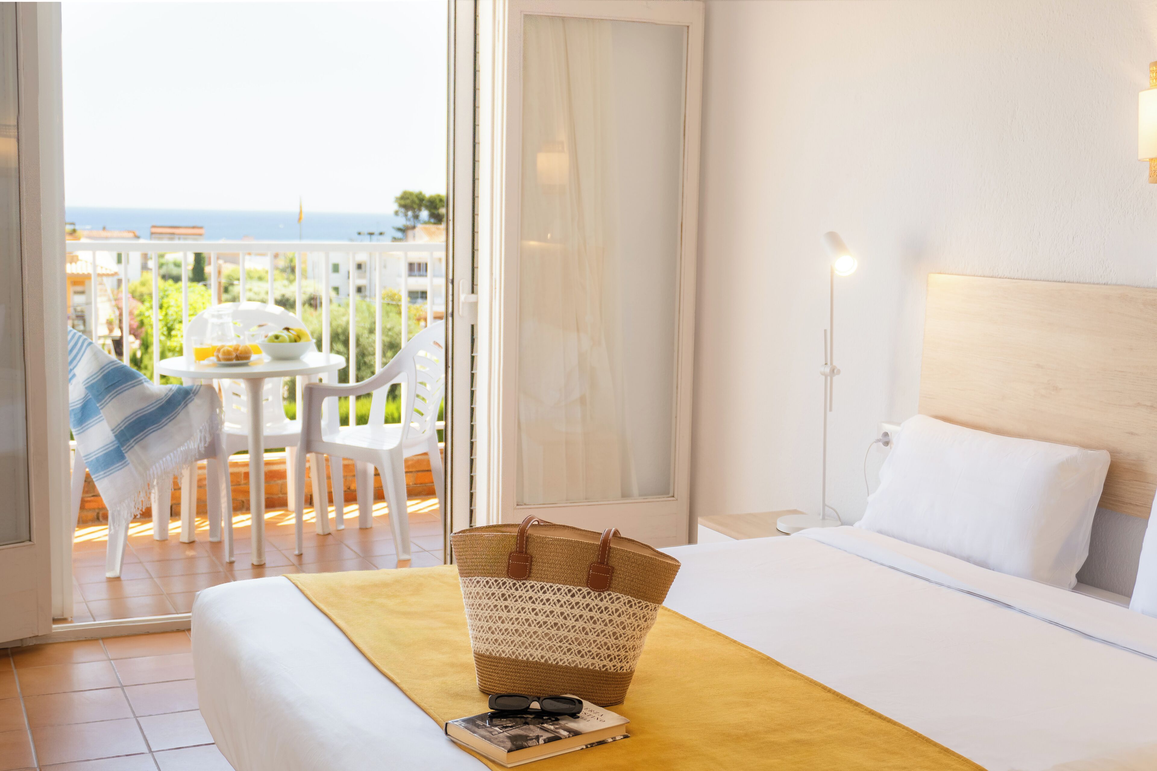 A hotel room with a bed, a bedside lamp, a window with a view of the sea, a balcony with chairs, and a table with breakfast items.