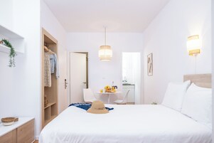 A hotel room with a bed, a wardrobe, a small table with a chair, and a kitchenette in the background.