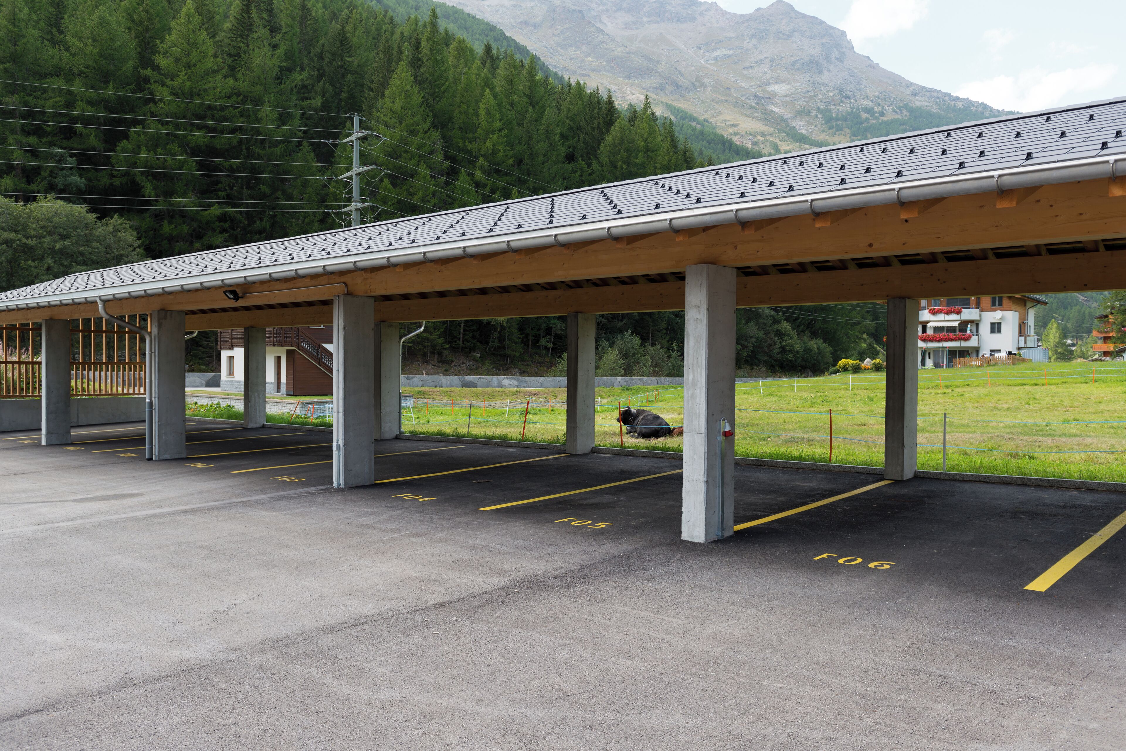 Een overdekte parkeerplaats met aangegeven parkeerplaatsen, een berg achtergrond en een gebouw met rode accenten in de verte.