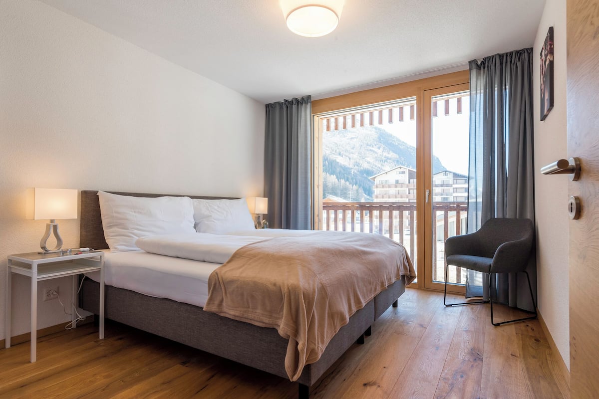 A bedroom with a bed, bedside table, chair, and a view of mountains through a balcony.