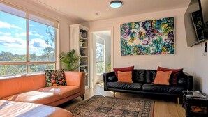 Een woonkamer met een leren bank, een zwarte bank met oranje kussens, een kleurrijk abstract schilderij en een groot raam met uitzicht op bomen.