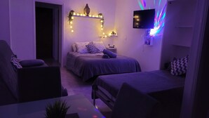 Une petite pièce faiblement éclairée, avec un lit, un canapé et un téléviseur fixé au mur.