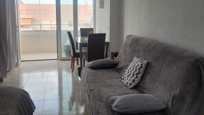 Un salon avec un canapé gris, une petite table et des chaises. Une porte-fenêtre coulissante donne accès à un balcon extérieur.