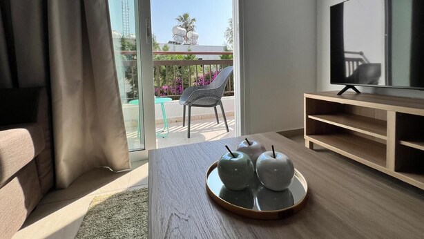 A modern living room with a sofa, a coffee table with decorative apples, a TV on a wooden stand, and a sliding glass door leading to an outdoor area with a chair and plants. - Cosy 1/bdr central apart near the beach

 (Paphos)