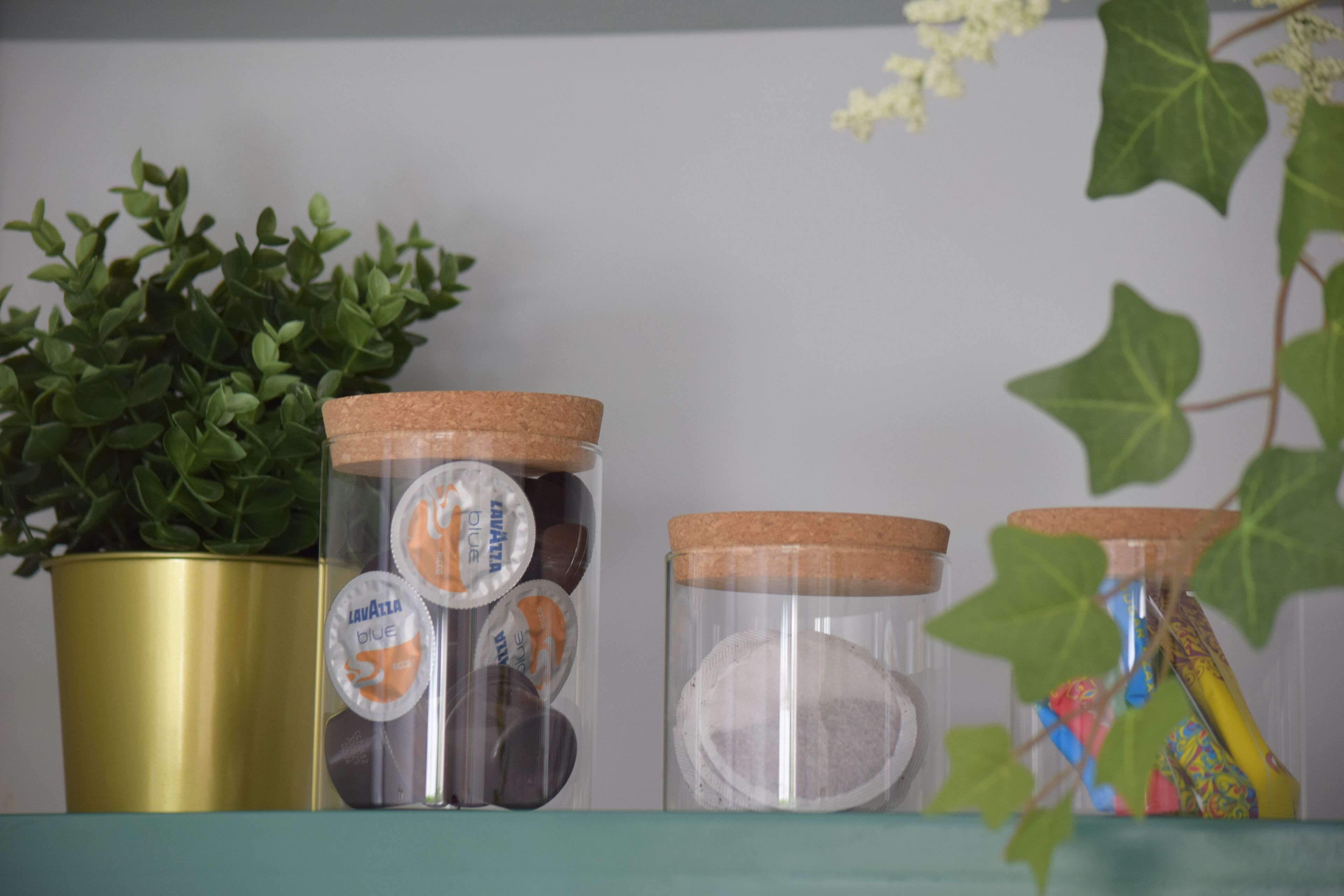 Three glass jars with cork lids on a shelf, one with a gold-colored pot beside it.