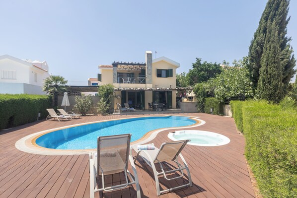 Ein Schwimmbecken mit Liegestühlen und einer Whirlpool, umgeben von einer Holzterrasse und Grünflächen.