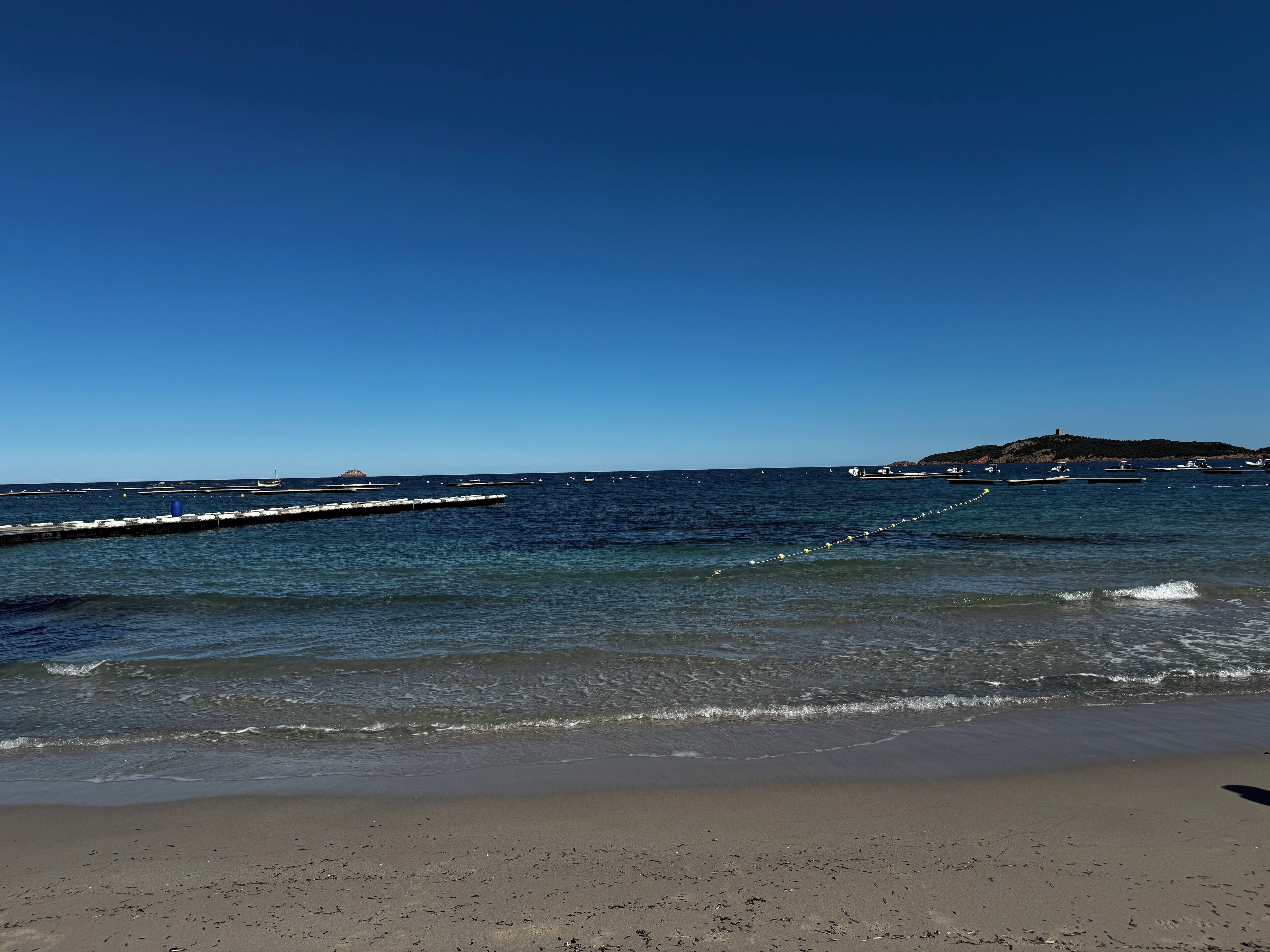 Une plage à l’eau d’un bleu cristallin, avec un rivage sablonneux et, au loin, une île parsemée de bâtiments.