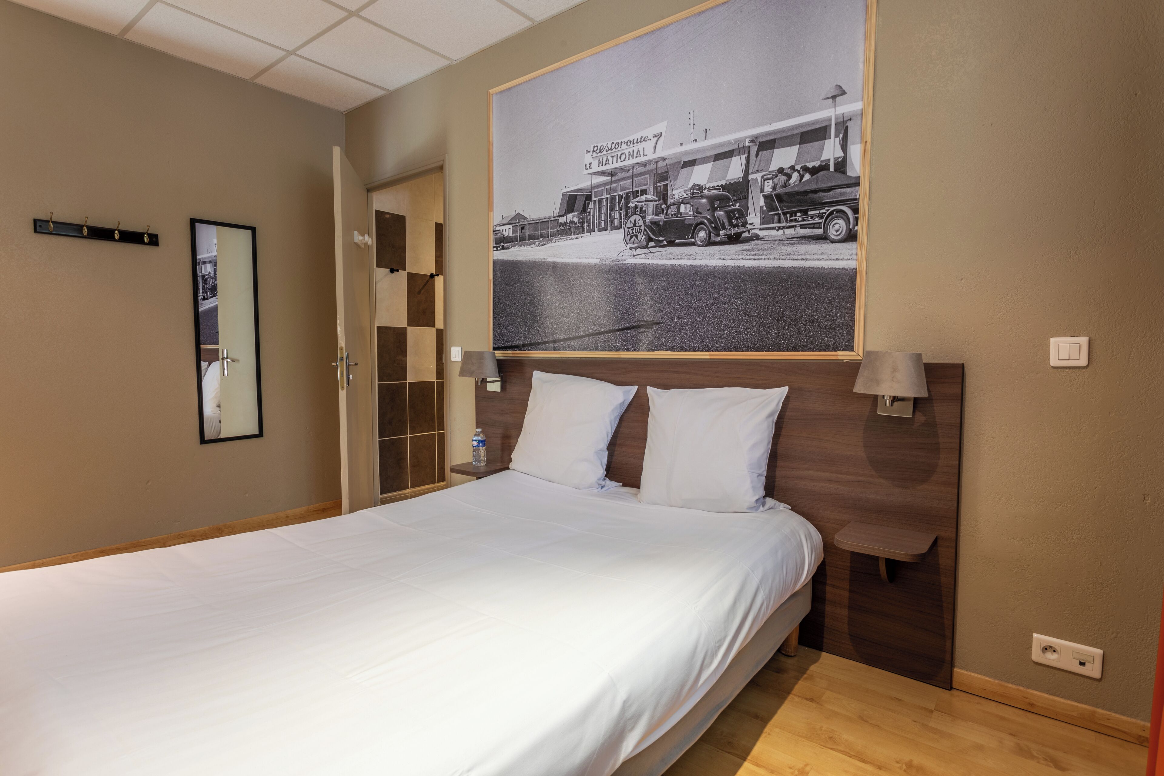 Una cama bien hecha en una habitación de hotel con un cuadro grande colgado encima, una mesita de noche con una lámpara y un estante pequeño.
