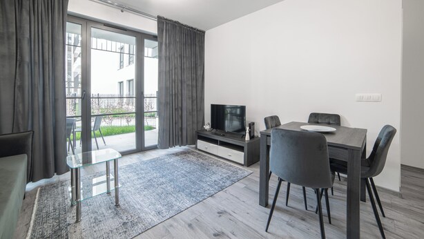 Ein modernes Wohnzimmer mit einem Glastisch, einem Esstisch mit Stühlen, einem Flachbildfernseher und einem großen Fenster mit Vorhängen.
