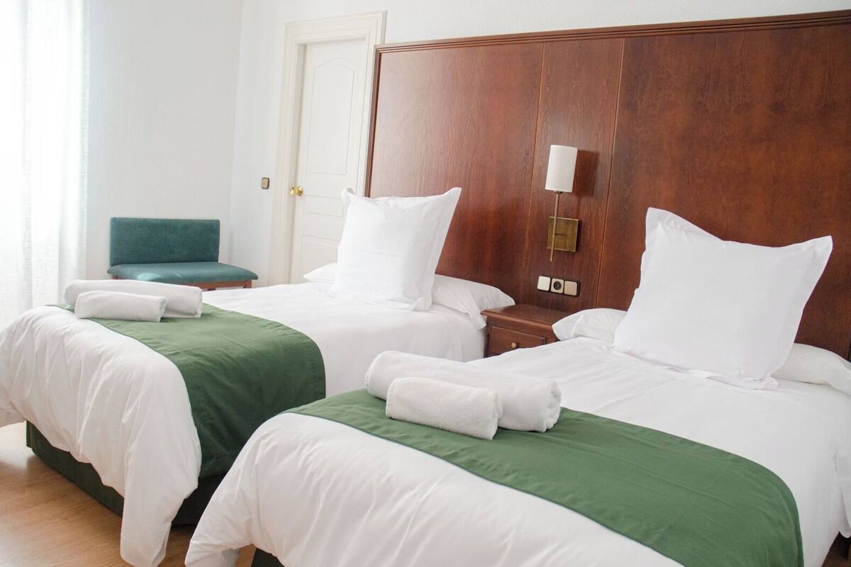 Habitación de hotel con dos camas, cada una con ropa blanca y cubrecamas verdes. Se observa un cabecero de madera, una mesita de noche con una lámpara, y una silla verde en la esquina.