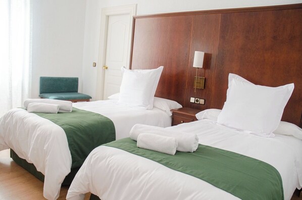 Une chambre d’hôtel avec deux lits, chacun agrémenté de draps blancs et d’une couvre-lit verte. On y trouve une tête de lit en bois, une table de chevet avec une lampe et une chaise verte dans un coin.