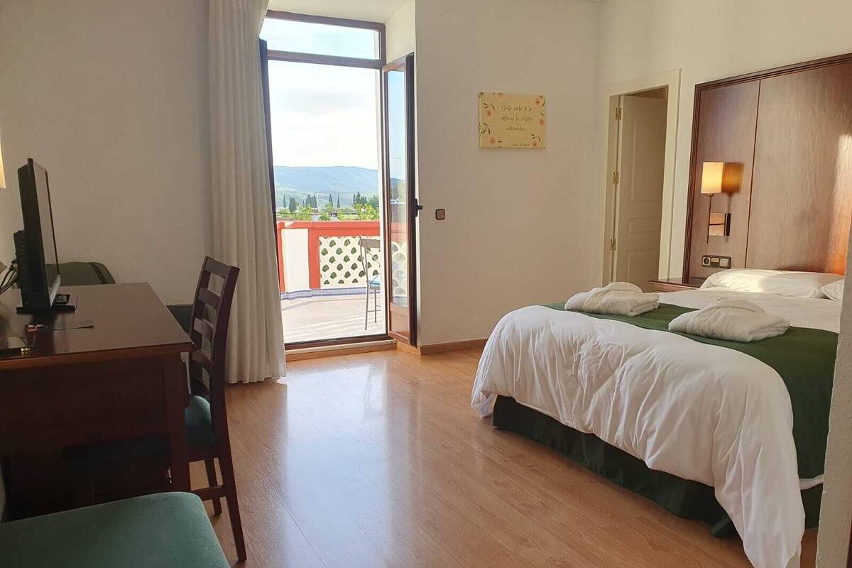 Habitación de hotel con escritorio, cama con ropa blanca, vista a través de una puerta de cristal y una lámpara fijada a la pared.