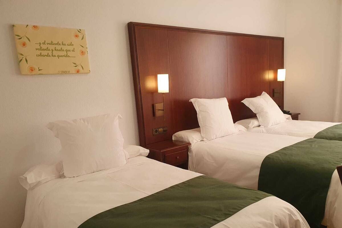 Habitación de hotel con dos camas, cada una con ropa blanca y colchas verdes. Cada cama tiene una cabecera de madera con una mesita de noche y una lámpara a cada lado. Se ve un letrero decorativo con texto en la pared.
