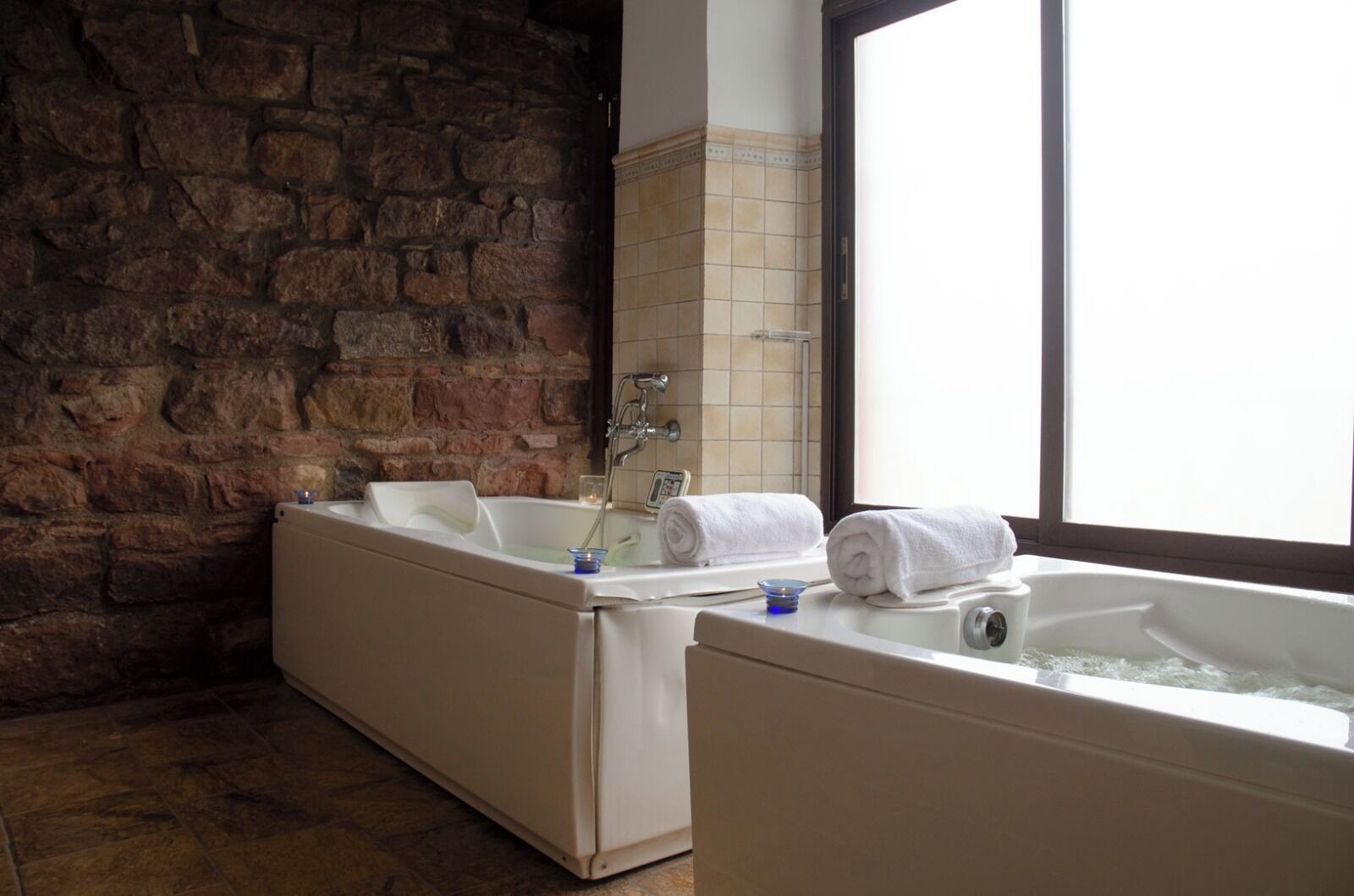 Un baño con dos bañeras, una pared de piedra y un ventanal grande.
