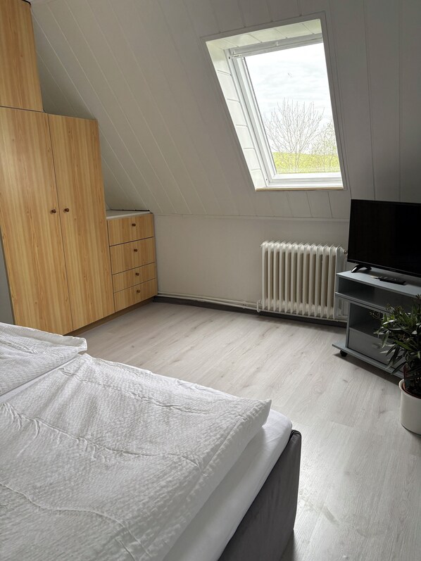 A bedroom with a sloped ceiling, a skylight, a wooden wardrobe, a radiator, a TV on a stand, and a potted plant. - Kleine Ferienwohnung , Direkt am Deich und Hafen von Eckwarden mit Gartennutzung (Butjadingen)