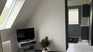 Una camera da letto con soffitto spiovente, una TV su un mobile, una pianta in vaso, un letto e vista su un balcone.
