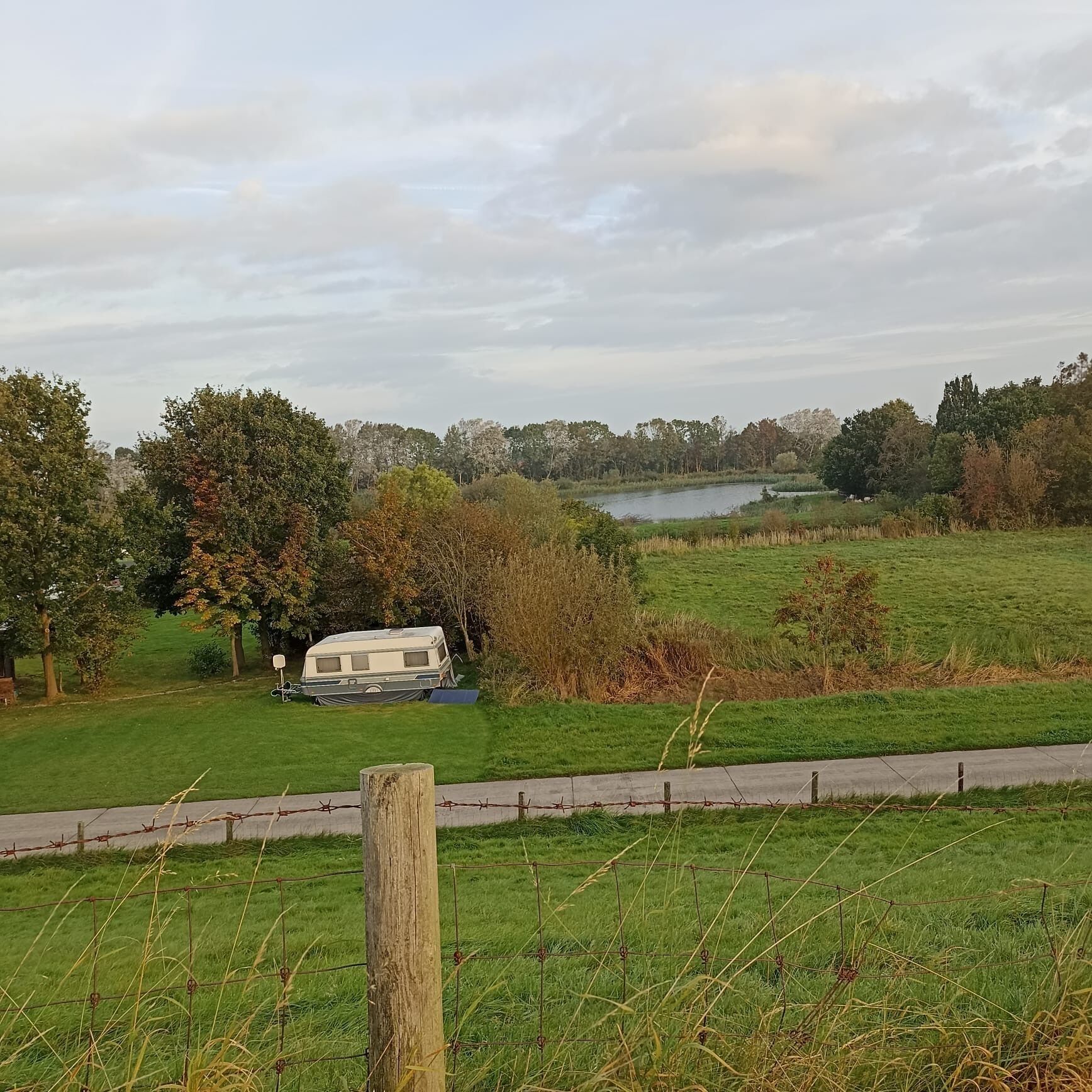 Un prato erboso con un lago, alberi e un camper parcheggiato vicino a una recinzione in legno.