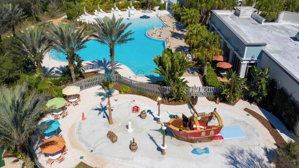An aerial view of a resort with a large swimming pool, palm trees, and a play area with a pirate ship.