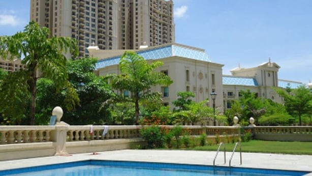 Piscina com água azul cristalina, rodeada por árvores verdes exuberantes e um edifício com fachada clara e telhado azul.