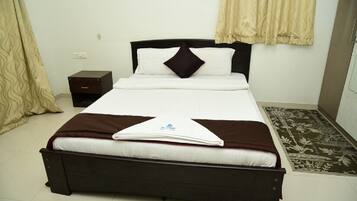 Una cama bien hecha con cabecera oscura y ropa de cama blanca, una mesita de noche pequeña y una alfombra estampada en el suelo.