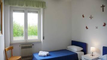 Ein Schlafzimmer mit einem Einzelbett, einem Sessel, einem Heizkörper, einem Fenster mit Blick auf Grünflächen und einer Wand mit Kreuz- und Schmetterlingsdekorationen.