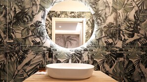 A bathroom with a round mirror, a white sink, and a wooden countertop. The walls are decorated with a tropical leaf pattern. - Vatican Cozy Suites (Rome)