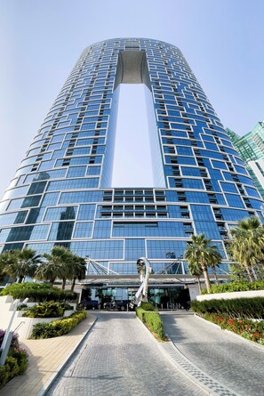 A modern high-rise building with a distinctive central atrium, surrounded by landscaped areas and palm trees. - Famous Address Beach Resort JBR 1BR (Dubai)