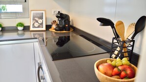 Une cuisine dotée d’une plaque de cuisson à induction noire, d’une machine à café, d’un saladier rempli de fruits et d’ustensiles de cuisine en bois posés sur le plan de travail.