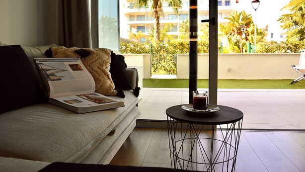 Un salon moderne avec un canapé, une table basse et une vue sur un balcon agrémenté de palmiers et d’un immeuble en arrière-plan.