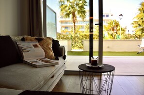 A modern living room with a sofa, a coffee table, and a view of a balcony with palm trees and a building in the background. - Villa Marina - Apartamento en Denia (Dénia)