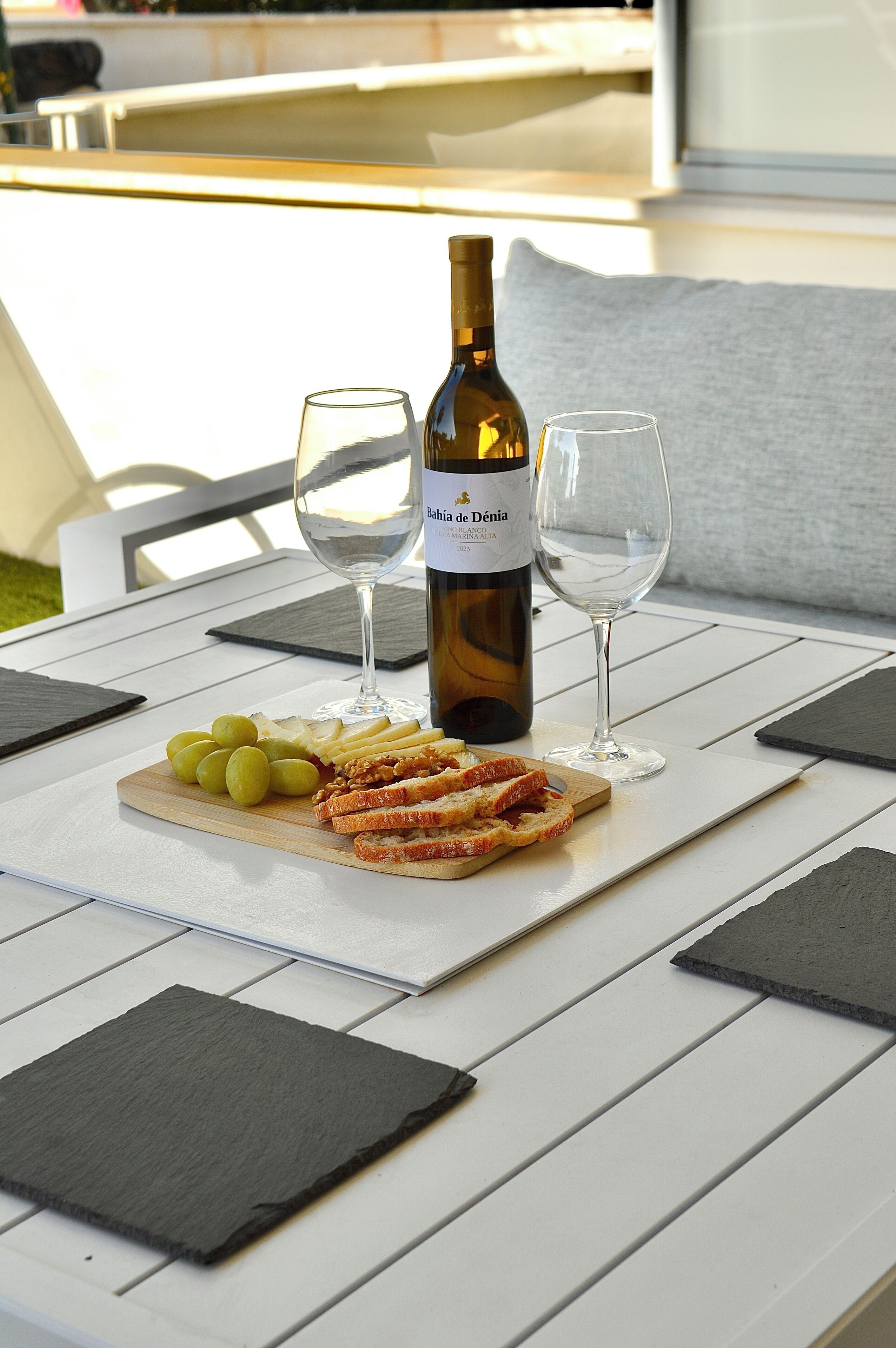Uma mesa com uma garrafa de vinho, duas taças, um prato com uvas e pão fatiado, e um jogo americano.