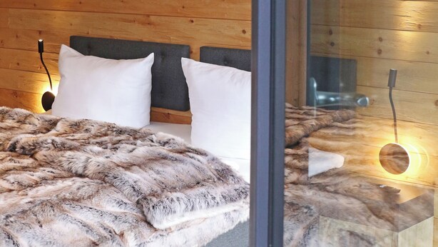 Un dormitorio moderno con separador de vidrio, paredes de madera, una cama con ropa de cama blanca y gris, y una mesita de noche con lámpara.