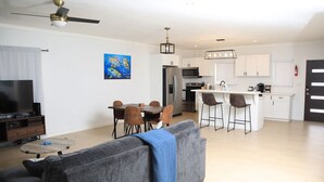 A modern living room with a grey sofa, a coffee table, and a TV. There is a dining area with a table and chairs, and a kitchen with white cabinets and appliances. - Townhouse w/Pool Near Beach, Airport & Supermarket (Nassau)