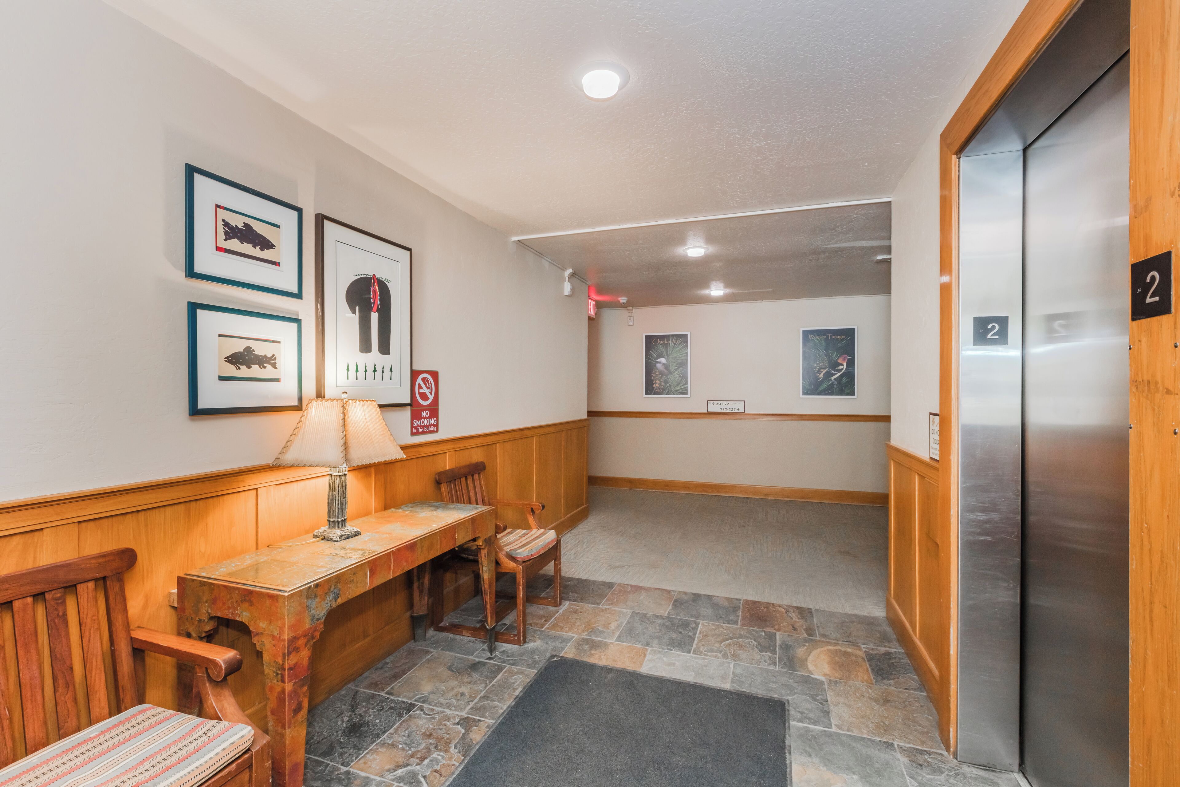 A lobby with a wooden bench, a table with a lamp, framed artwork on the wall, and an elevator.