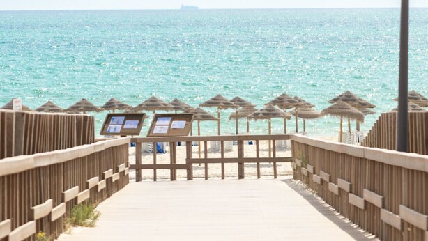Un passeig de fusta que condueix a una platja amb paraigües de palla i cartells informatius.