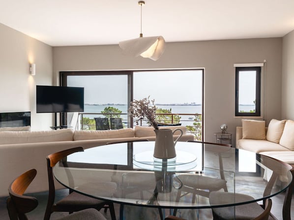 A modern living room with a glass dining table, sofas, a TV, and large windows offering a view of the water.