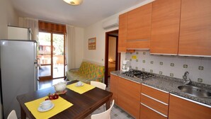 A compact kitchen with wooden cabinets, a gas stove, and a sink. There is a dining area with a table set for two and a colorful sofa. - Family escape in Bibione (Bibione)
