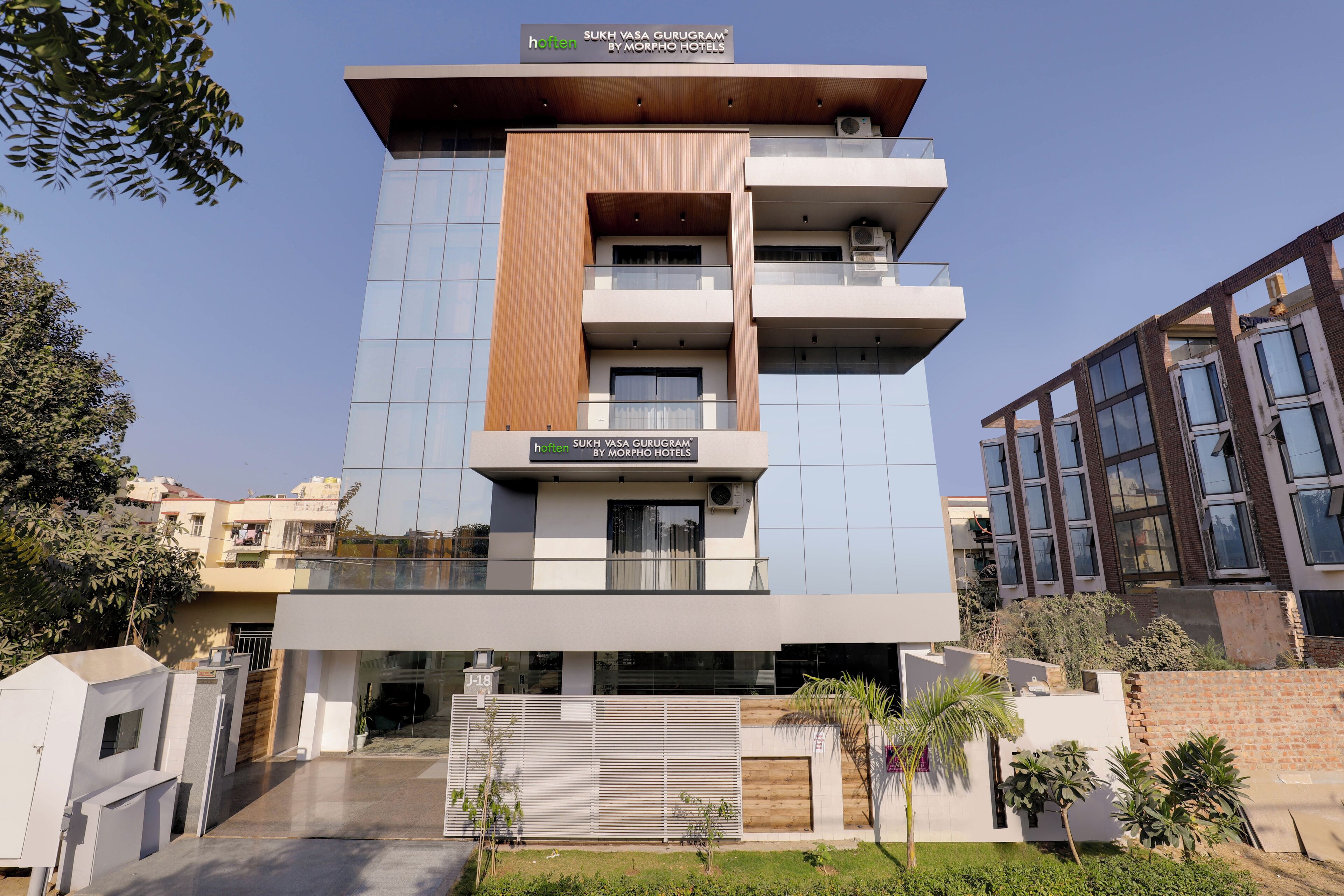 A modern hotel building with a sign reading "Sukh Vasa Gurugram by Morpho Hotels".