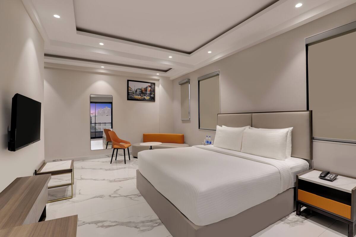 Une chambre d’hôtel moderne avec un grand lit, un bureau et une chaise. Un téléviseur à écran plat est fixé au mur, il y a une fenêtre donnant sur la ville et une porte avec un miroir.