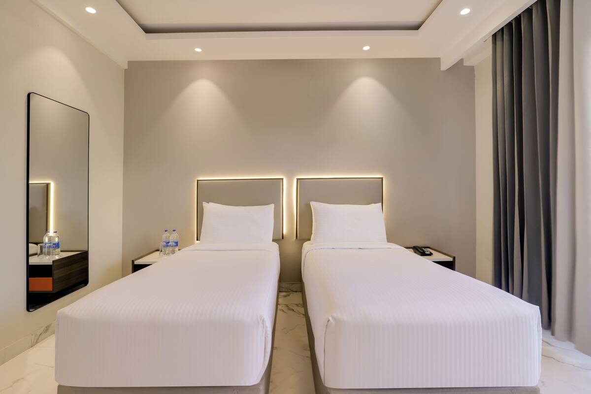 Une chambre d’hôtel avec deux lits simples, un grand miroir, des tables de chevet avec des bouteilles d’eau et un téléphone.