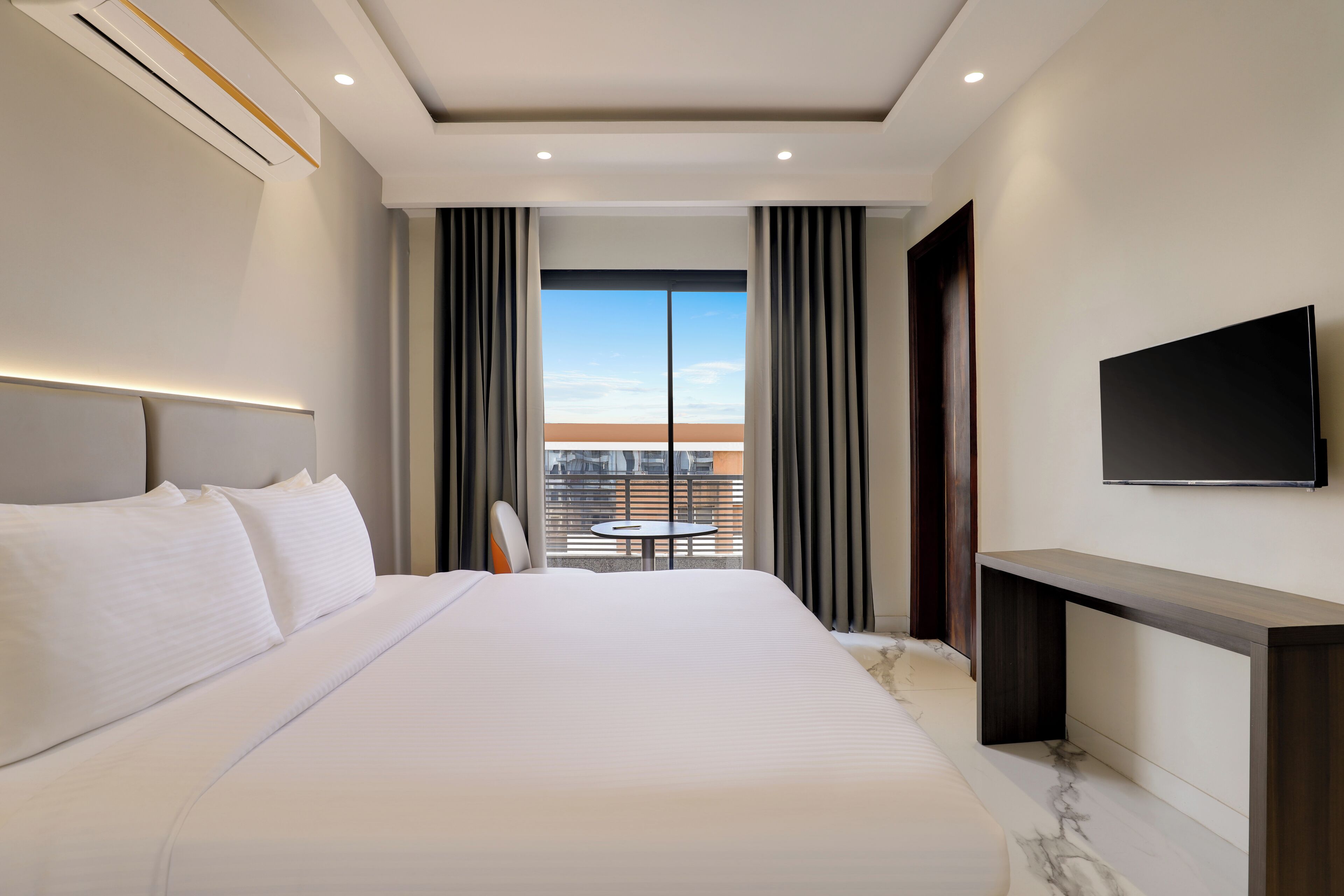 A modern hotel room with a large bed, a TV, a balcony with a table, and a city view.